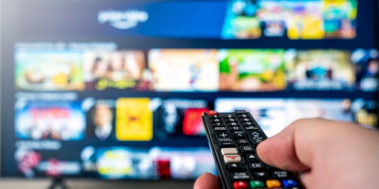 Hand with tv remote aimed towards big 4k flat screen oled tv with blurred out tv entertainment system meny. Technology and quarantine concept.